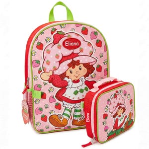 May include: A pink backpack and matching lunch bag set featuring a Strawberry Shortcake character design. The backpack has a large main compartment, side pocket, and green handle. The lunch bag has a zipper closure and a top handle. Both items are decorated with strawberries.