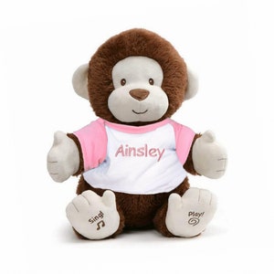 May include: Brown plush monkey wearing a white t-shirt with pink sleeves and the name "Ainsley" embroidered on the front. The monkey has a brown face and brown fur. The monkey's feet have the words "Sing!" and "Play!" embroidered on them.