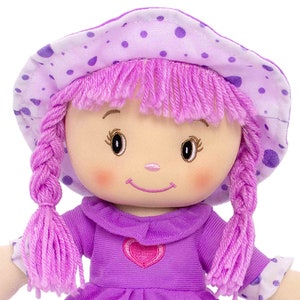 Peut inclure: Une poup&eacute;e en peluche douce, v&ecirc;tue d'une robe violet clair et d'un chapeau assorti &agrave; pois violets fonc&eacute;s. La poup&eacute;e a des cheveux en fil rose tress&eacute;s et un visage souriant avec des joues roses. Un c&oelig;ur rose est brod&eacute; sur la robe.