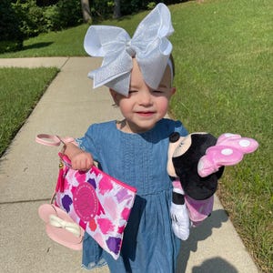 May include: A young girl wearing a blue denim dress and a silver glitter bow headband holds a pink and white purse with a pink Minnie Mouse head charm. She is also holding a pink and black Minnie Mouse plush toy.