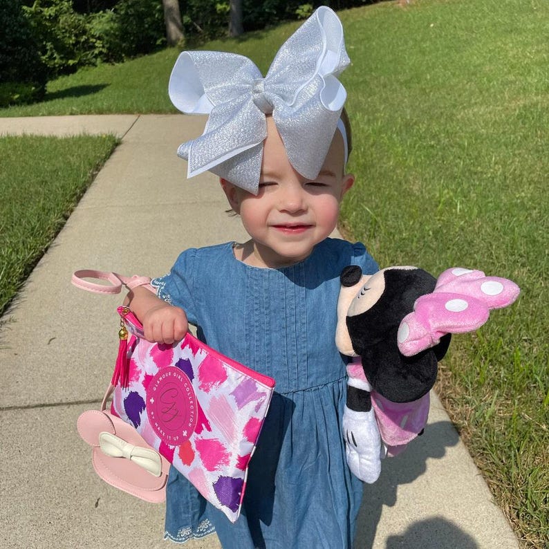 May include: A young girl wearing a blue denim dress and a silver glitter bow headband holds a pink and white purse with a pink Minnie Mouse head charm. She is also holding a pink and black Minnie Mouse plush toy.