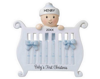 May include: White baby crib ornament with a blue and white pattern. A baby with a blue hat is peeking over the top of the crib. The ornament says "HENRY 20XX Baby's First Christmas".