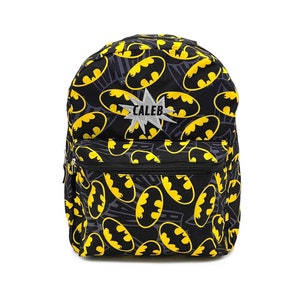 May include: Black backpack with a repeating pattern of yellow Batman logos and gray accents. The backpack has a front zippered pocket and a silver starburst patch with the name "CALEB" in white letters.