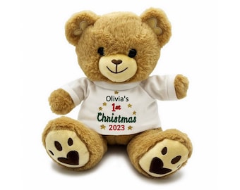 May include: A brown teddy bear wearing a white shirt with the text "Olivia's 1st Christmas 2023" embroidered on it. The bear has paw prints on its feet.