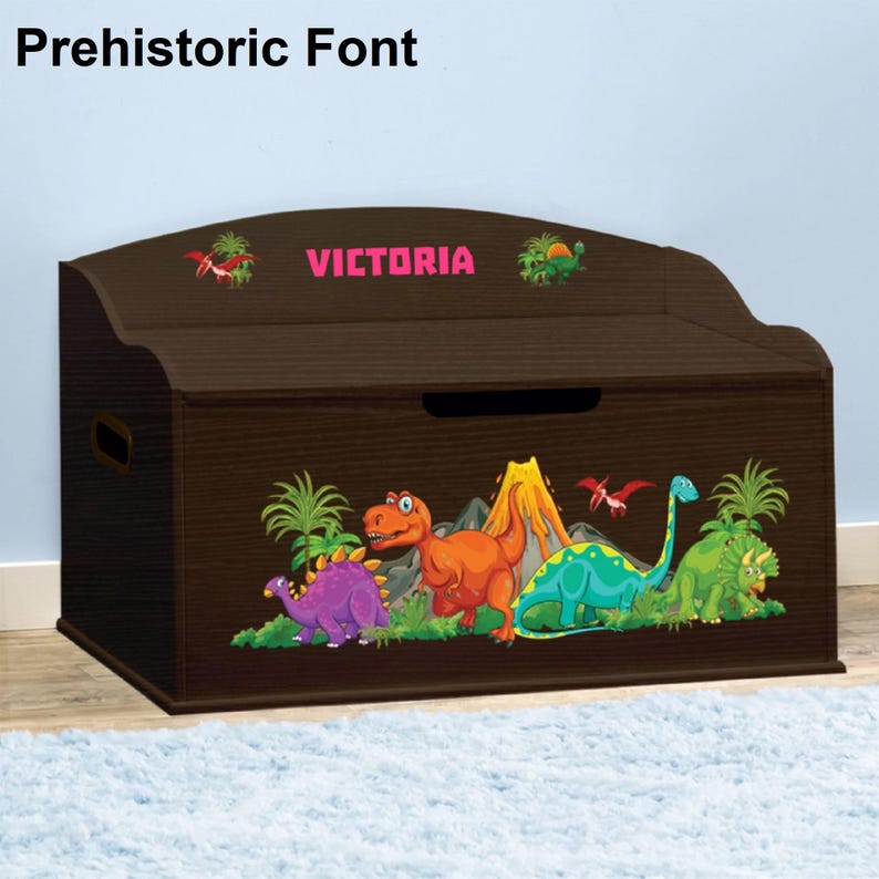 May include: A brown wooden toy chest with a dinosaur theme. The chest has a hinged lid and a handle on the side. The front of the chest features a colorful graphic of dinosaurs, a volcano, and palm trees. The name "VICTORIA" is printed in pink letters on the top of the chest.
