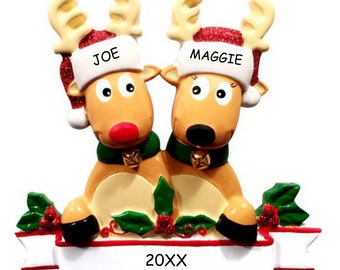 May include: Personalized Christmas ornament featuring a couple of reindeer wearing Santa hats with the names "JOE" and "MAGGIE" on them. The reindeer are sitting together on a banner with the year "20XX" printed on it.