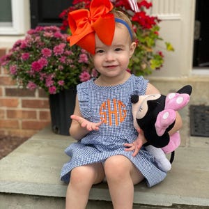 May include: A young girl wearing a blue and white gingham dress with an orange monogrammed "SHG" on the front. She is holding a pink and black plush toy and wearing a large orange bow in her hair.