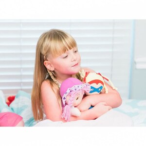 Peut inclure: Une jeune fille aux longs cheveux blonds tient deux poup&eacute;es en peluche. L'une a un chapeau rose &agrave; pois blancs, l'autre est une poup&eacute;e sur le th&egrave;me des super-h&eacute;ros avec un design rouge et bleu. Les poup&eacute;es sont douces et c&acirc;lines.