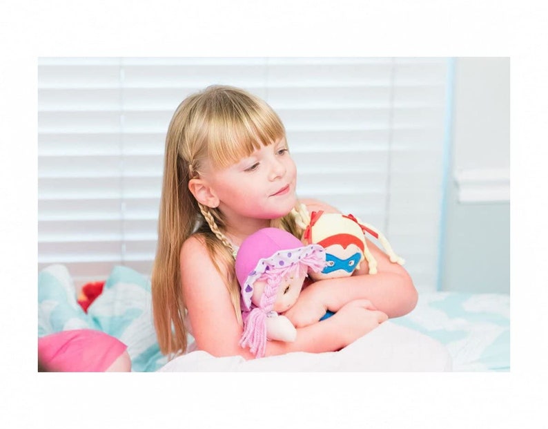 Peut inclure: Une jeune fille aux longs cheveux blonds tient deux poup&eacute;es en peluche. L'une a un chapeau rose &agrave; pois blancs, l'autre est une poup&eacute;e sur le th&egrave;me des super-h&eacute;ros avec un design rouge et bleu. Les poup&eacute;es sont douces et c&acirc;lines.