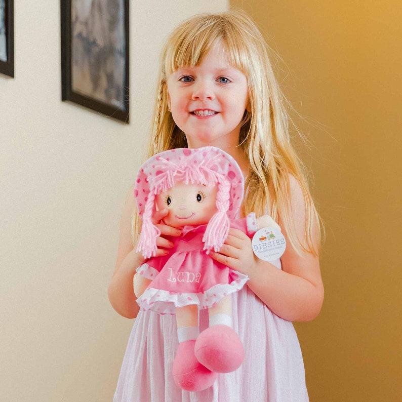Peut inclure: Une poup&eacute;e de chiffon rose avec un chapeau, une robe et des chaussures assortis. La poup&eacute;e a des cheveux blonds en fil et le nom "Luna" brod&eacute; sur la robe. La poup&eacute;e est tenue par une jeune fille aux cheveux blonds.