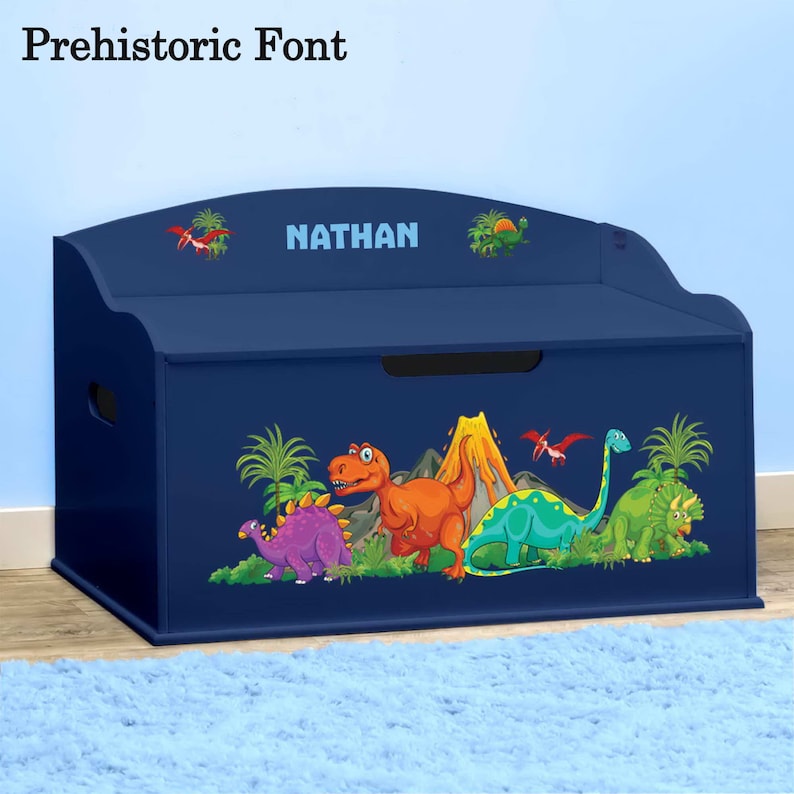 May include: A blue wooden toy chest with a dinosaur theme. The chest has a hinged lid and a handle on the side. The front of the chest features a colourful illustration of dinosaurs, a volcano, and palm trees. The name "NATHAN" is printed on the top of the chest.