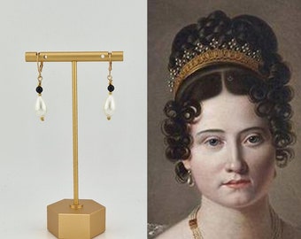 May include: Gold-tone leverback earrings with a teardrop pearl and black bead accent. The earrings are displayed on a gold stand next to a portrait of a woman with a pearl earring. The earrings are a classic jewelry design.