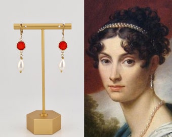 May include: A pair of gold dangle earrings with red glass beads and white pearl drops. The earrings are displayed on a gold stand.
