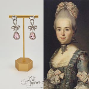 May include: A pair of silver dangle earrings with pink teardrop crystals and a bow detail. The earrings are displayed on a gold stand.
