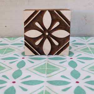May include: A wooden block printing stamp with a carved floral design. The stamp is brown with a white floral pattern in the centre. The stamp is sitting on a white fabric with a green and red geometric pattern.