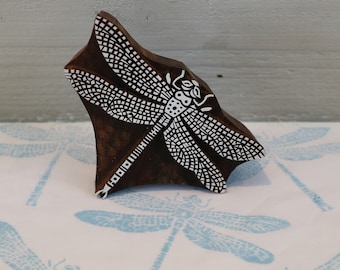 May include: A wooden block carving of a dragonfly with white details. The carving is used as a stamp for creating patterns on fabric.