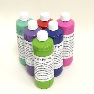 Op de afbeelding: Zes flessen ambachtelijke stoffenverf in verschillende kleuren, waaronder groen, paars, rood, roze en blauw. De flessen zijn gelabeld met "Craft Fabric Paint" en hebben witte doppen.
