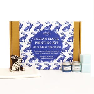 May include: An Indian block printing kit for a hare and star tea towel, featuring a cardboard box with a blue and white hare pattern, a wooden hare printing block, a small star block, two jars of fabric paint, and a plain white cotton tea towel.