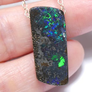 May include: A rectangular pendant featuring a dark, textured boulder opal with vibrant flashes of neon green and deep blue color play. The stone is suspended from a delicate, thin silver chain, showcasing a natural, raw surface with visible cracks.