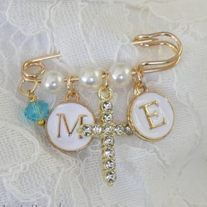 May include: Gold-toned safety pin with pearl beads, a cross pendant encrusted with rhinestones, and two white enamel charms with the letters "M" and "E". A blue bead dangles from the pin. The pin is set against a white lace background.