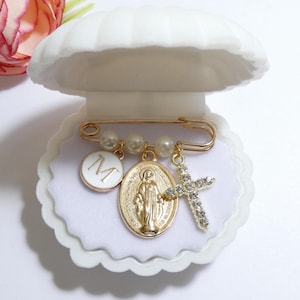 May include: A gold-tone safety pin brooch featuring three white faux pearls, a circular white charm with the letter M, an oval gold-tone medal depicting a religious figure, and a cross charm encrusted with clear crystals inside a white shell case.