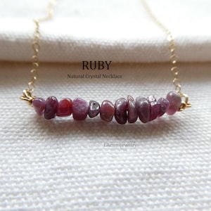 May include: A necklace featuring a row of small, raw, deep pinkish-red ruby crystal chips strung on a thin gold-toned metal chain. The text RUBY Natural Crystal Necklace and LAminiJewelry are visible in the center and bottom of the image.