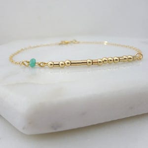 May include: A delicate gold bracelet with small, round beads and cylindrical bars. A single, small, light-green bead is set near the clasp. The bracelet is displayed on a white marble surface.