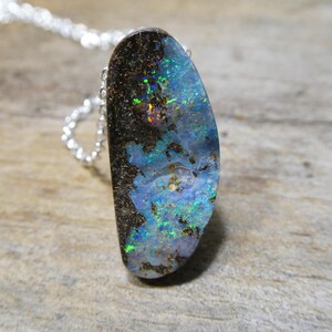 May include: A pendant necklace featuring an elongated, irregular dark boulder opal with vibrant flashes of blue, green, and orange fire. The stone is set in a simple metal frame and hangs from a delicate, thin silver-toned link chain.