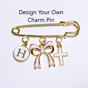 May include: A gold-toned safety pin brooch featuring three dangling charms: a white circular charm with the letter H, a light pink bow charm, and a white cross charm. The text Design Your Own Charm Pin is printed above the gold safety pin.
