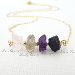 May include: A gold chain necklace featuring four raw, uncut gemstones arranged horizontally: a pale pink rose quartz, a translucent smoky quartz, a deep purple amethyst, and a dark black tourmaline, with text labels identifying each stone.