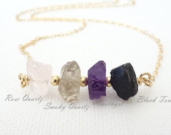 May include: A gold chain necklace featuring four raw, uncut gemstones arranged horizontally: a pale pink rose quartz, a translucent smoky quartz, a deep purple amethyst, and a dark black tourmaline, with text labels identifying each stone.