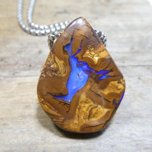 May include: A teardrop-shaped boulder opal pendant featuring a polished brown ironstone matrix with deep, vibrant blue flashes of colour throughout the stone. The pendant is suspended from a silver-toned metal ball chain necklace.