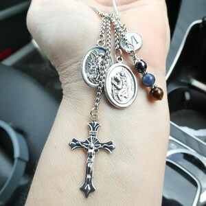 May include: A silver-toned car charm featuring a crucifix, religious medallions, and a charm with the letter "V". The charm also includes a strand of beads in black, blue, and brown. The charm is suspended from a silver chain.