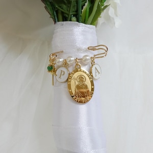 May include: A gold brooch with a safety pin design, featuring charms. These include a green gem, white lettered pendants, and a central oval pendant with an image of Saint Patrick. The brooch is attached to a white ribbon wrapped around a bouquet of flowers.