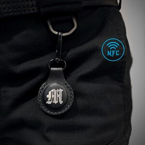 Könnte beinhalten: Schwarzer Leder-AirTag-Halter mit silbernem Initial "M" und schwarzem Karabinerhaken. Der Halter ist an einem schwarzen Gürtel befestigt. Der Text "LITTLE DESPOT LEATHERWORKS" ist auf dem Gürtel zu sehen. Ein blaues NFC-Symbol befindet sich auf der Hose.