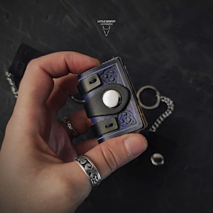 May include: A miniature blue and black leather book with Celtic knotwork and a silver snap closure. The small book is held in a hand and attached to a keychain. The logo "LITTLE DESPOT LEATHERWORKS" is visible in the upper left corner.