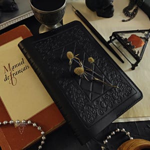 Puede incluir: Un diario de cuero negro con patrones florales y geométricos en relieve. El diario está adornado con flores secas y descansa junto a un libro antiguo titulado "Manuel de français". También se ven un bolígrafo, una caja de cristal decorativa y una copa de plata.