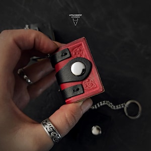 May include: A small, red leather book-shaped keychain with black accents and Celtic knot designs. The keychain has a silver-colored metal clasp and a chain. The text "LITTLE DESPOT LEATHERWORKS" is visible.