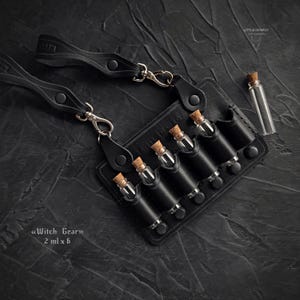 Könnte beinhalten: Schwarzer Leder-Fläschchenhalter mit sechs kleinen Glasflaschen, die jeweils mit einem Korken verschlossen sind. Der Halter hat Riemen mit Metallverschlüssen. Der Text "Witch Gear" und "2 ml x 6" ist sichtbar.