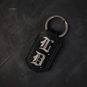 May include: Black leather keychain with silver-colored metal key ring. The keychain has a rectangular shape with rounded corners and features the initials "L.D." in a gothic font. The keychain is set against a dark background.