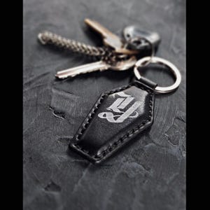 以下が含まれることがあります： 銀色の文字「J」のデザインが施された、黒い革製の棺型キーホルダー。 キーホルダーはシルバーのキーリングに付けられており、いくつかの鍵の隣にあります。 キーホルダーは縁が縫い付けられています。