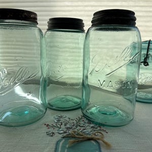 May include: Four vintage blue-green Ball Mason jars with black lids. The jars are of varying sizes and shapes, with embossed lettering. One jar has a wire closure. The jars are arranged on a white and blue patterned surface.