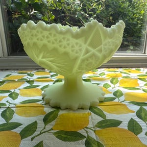 May include: A pale yellow, translucent glass pedestal bowl featuring a scalloped rim and intricate pressed geometric patterns, including star and diamond motifs, set on a circular base with a matching scalloped edge detail.