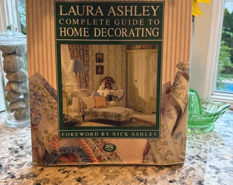May include: A hardcover book titled "LAURA ASHLEY COMPLETE GUIDE TO HOME DECORATING" with a floral design. The book is displayed on a countertop with sunflowers in the background. The cover features a room interior.