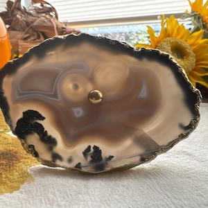 May include: A large, natural agate slice with a gold-toned metal rim and a small gold knob in the center. The agate has a light brown and white banded pattern with black inclusions.