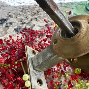 May include: Close-up of a vintage door handle with a tarnished brass finish. A metal tool is inserted into the handle. The handle is surrounded by red and green decorative beads, suggesting a holiday theme.