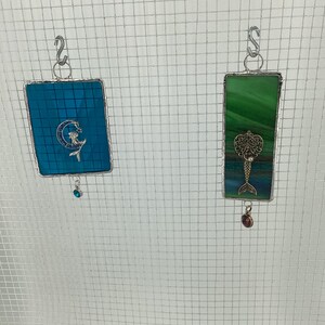 May include: Two stained glass suncatchers hanging from a wire mesh. The left suncatcher is blue with a mermaid design and a blue crystal hanging from the bottom. The right suncatcher is green with a mermaid design and a red crystal hanging from the bottom.