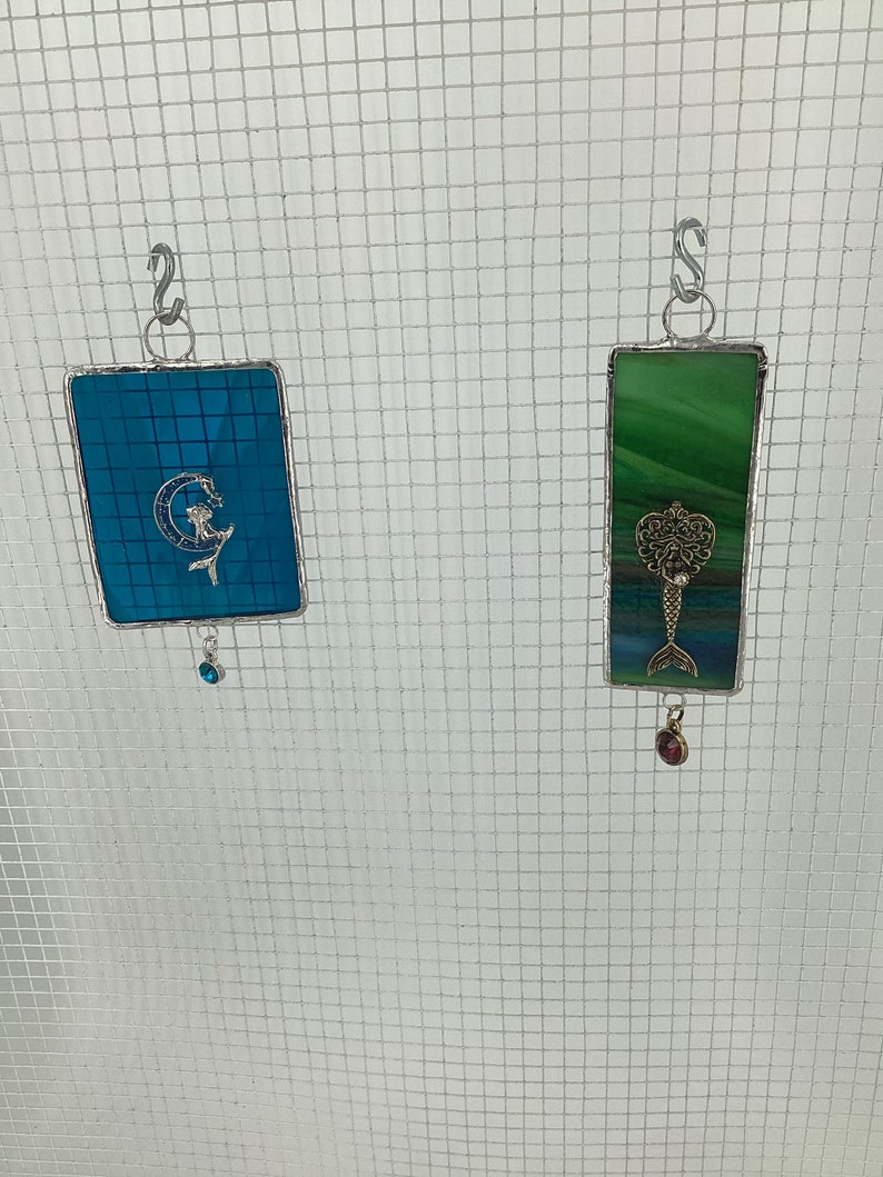 May include: Two stained glass suncatchers hanging from a wire mesh. The left suncatcher is blue with a mermaid design and a blue crystal hanging from the bottom. The right suncatcher is green with a mermaid design and a red crystal hanging from the bottom.