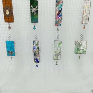 May include: Eight colourful glass ornaments with metal hangers and decorative charms. The ornaments are rectangular and feature various designs, including a fairy, a butterfly, and abstract patterns.