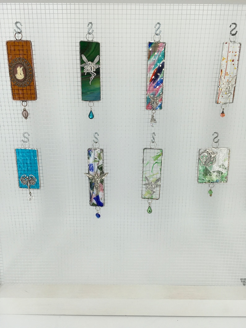May include: Eight colourful glass ornaments with metal hangers and decorative charms. The ornaments are rectangular and feature various designs, including a fairy, a butterfly, and abstract patterns.